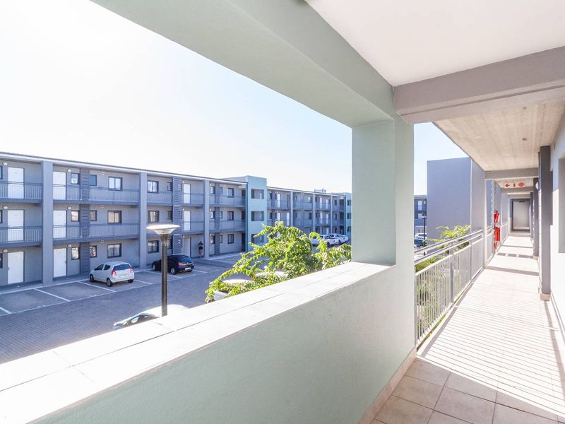 A covered balcony with tiled flooring and metal balustrades, overlooking the apartment complex parking area and building exterior.