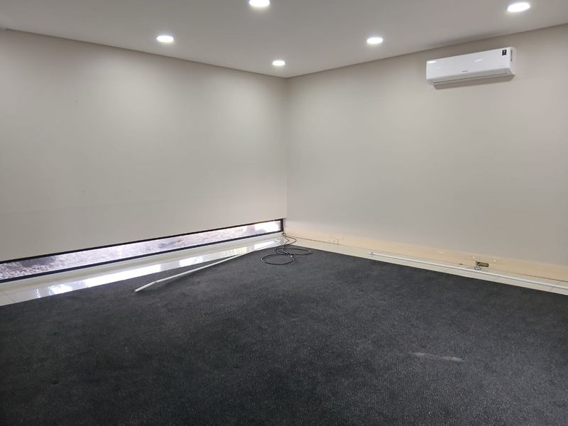A clean, empty office space featuring dark carpeting, recessed ceiling lighting, a split-system air conditioner, and a low-profile horizontal window.