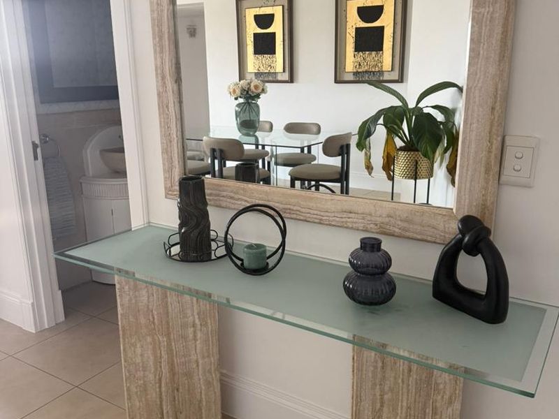 A bright entrance hall featuring a modern glass and travertine console table, large wall mirror, and neutral ceramic floor tiles.