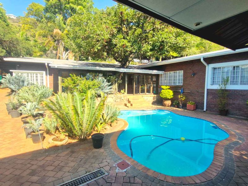 A private outdoor pool area featuring brick paving, established garden landscaping, and a covered patio section adjacent to the main brick dwelling.
