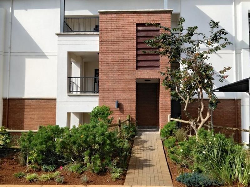 Front elevation of a modern apartment building featuring a brick facade, pitched roof detail, and a paved entrance walkway surrounded by landscaped garden beds.