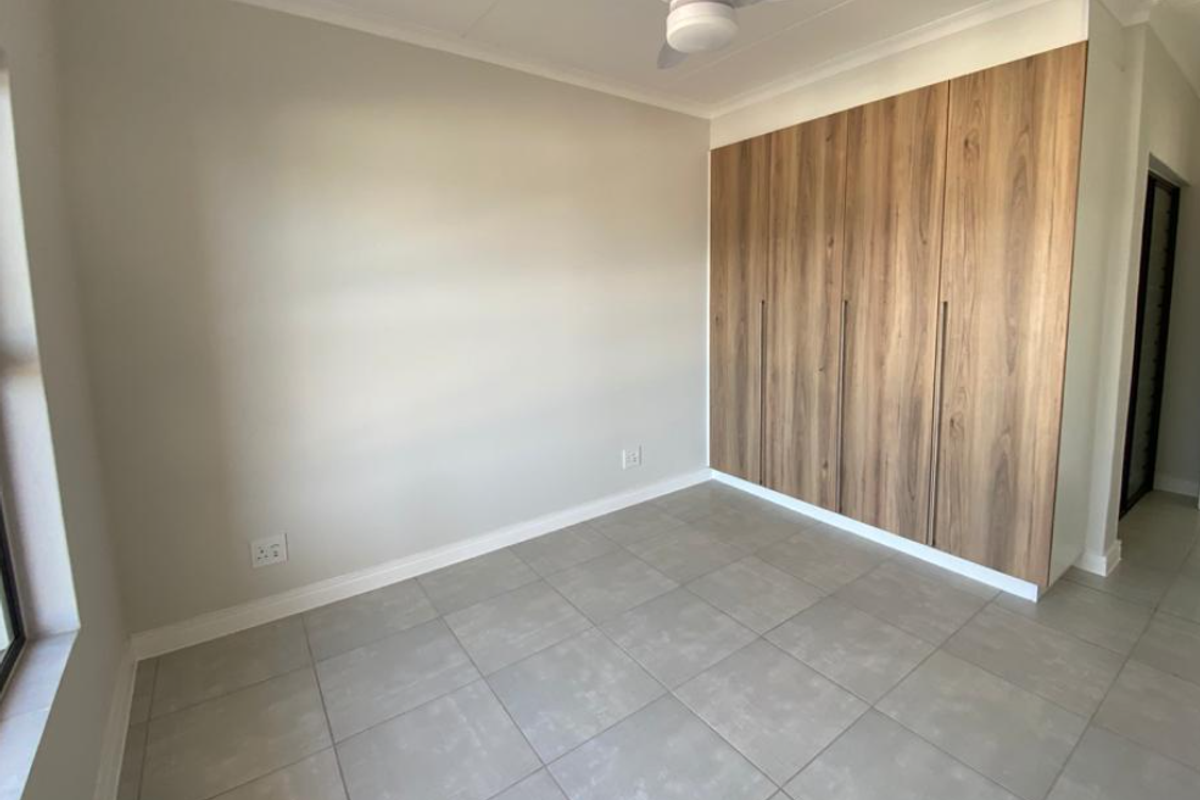 A bright, empty bedroom featuring modern wood-finish built-in cupboards, neutral walls, tiled flooring, and a ceiling fan.