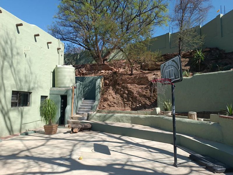Rear exterior area featuring a paved patio, concrete retaining walls, stairs, and a water storage tank set against a natural rock embankment.