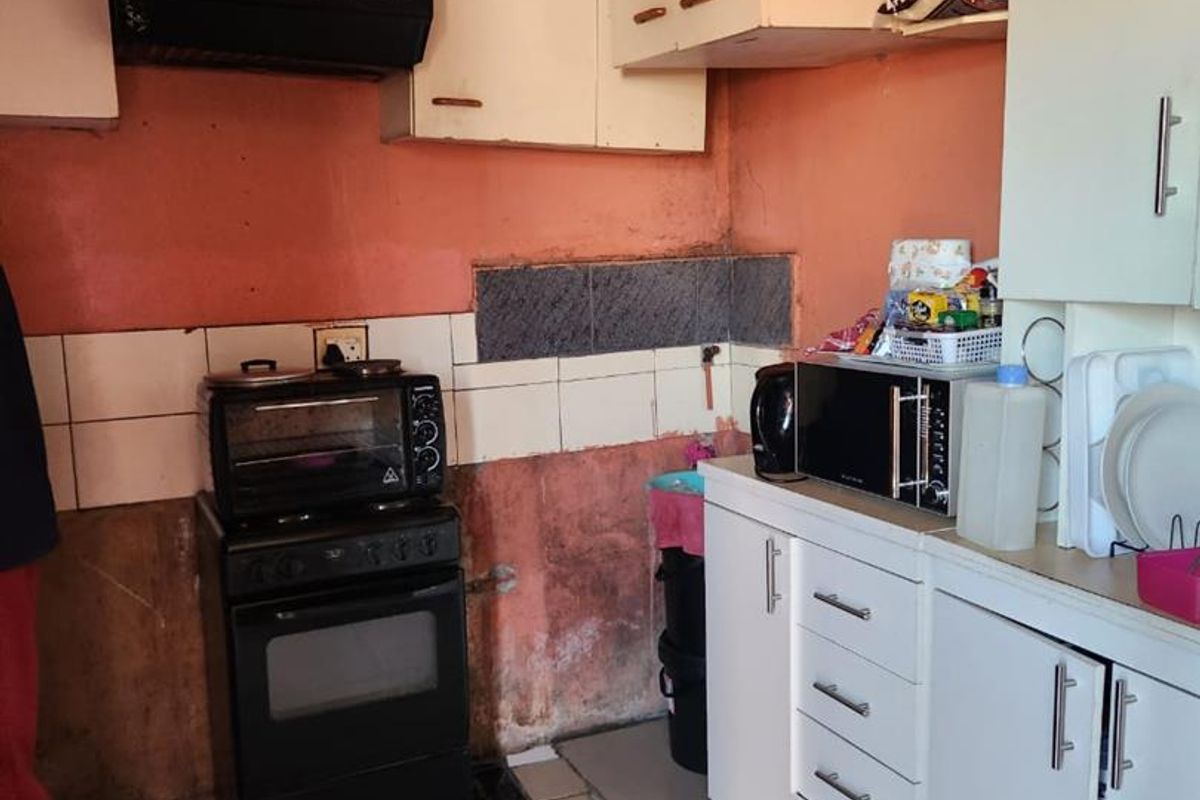 A dated kitchen featuring white cabinetry, a freestanding stove with extractor fan, and tiled flooring, requiring significant cosmetic updates and wall repairs.