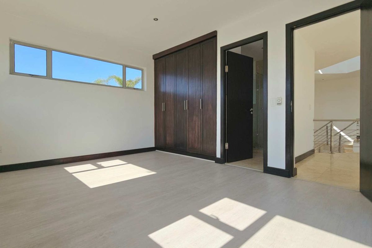 A bright, empty bedroom featuring modern laminate flooring, dark wood built-in cupboards, and direct access to an en-suite bathroom and hallway.