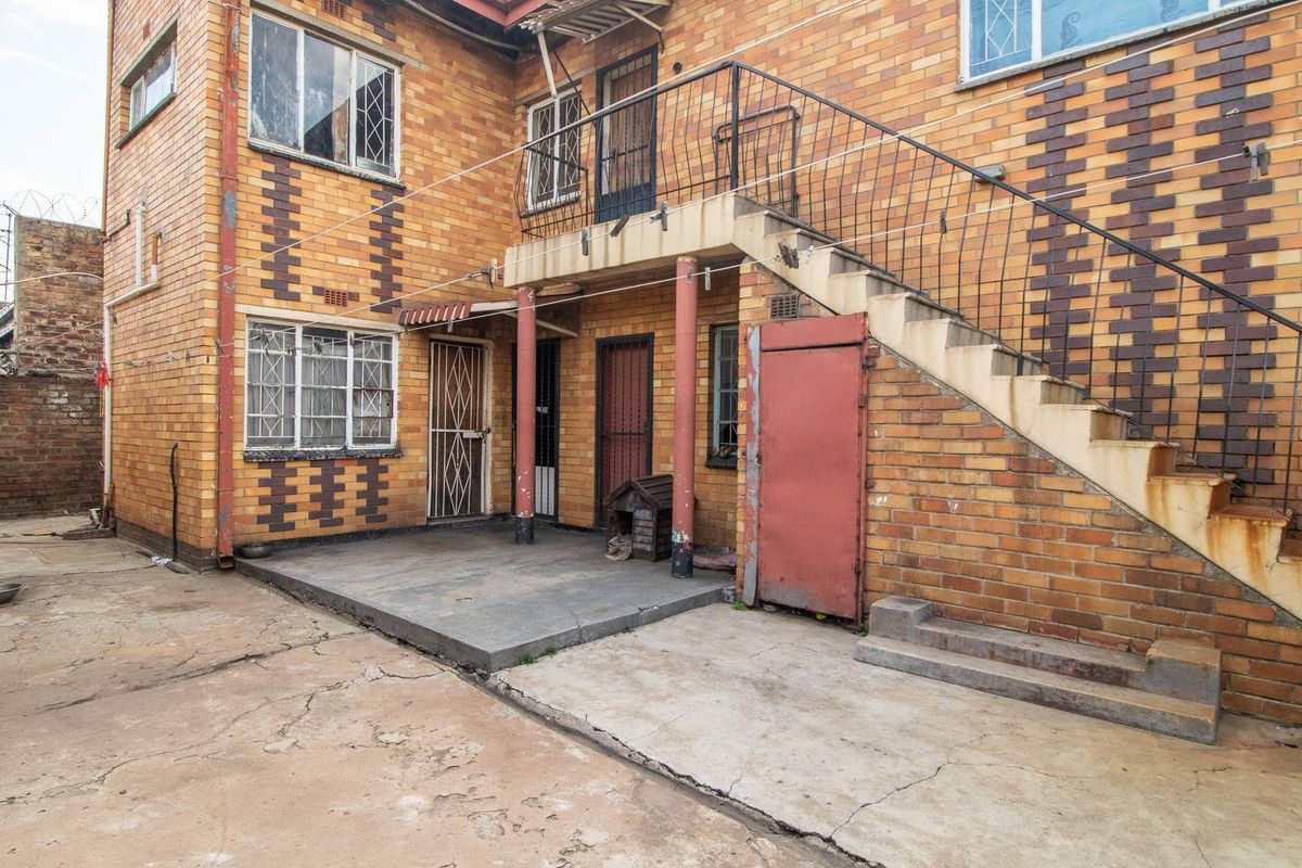 Rear exterior view of a multi-story brick building featuring an external concrete staircase, metal security gates, and a paved courtyard area.