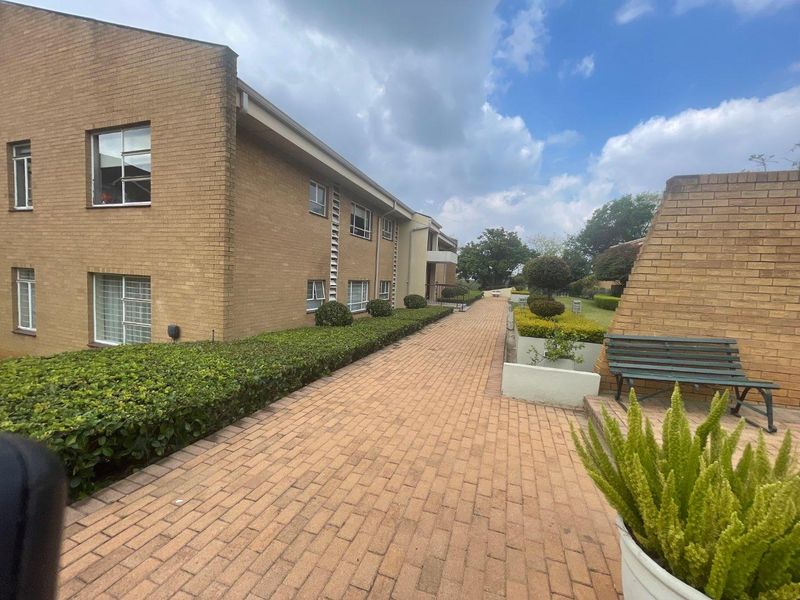 A well-maintained exterior view of the apartment complex featuring a paved walkway, brick facade, and neatly landscaped garden hedges.
