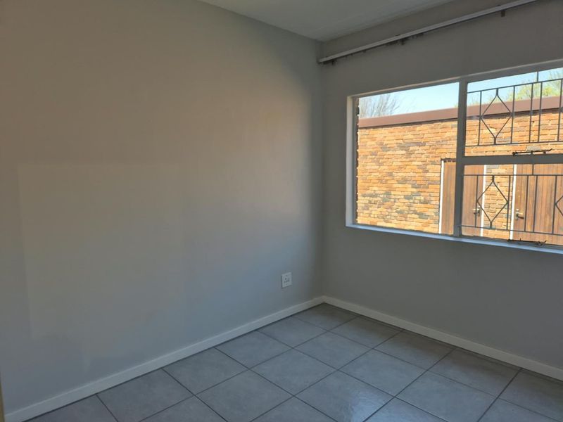 A clean, empty bedroom featuring neutral grey walls, tiled flooring, and a window fitted with security burglar bars.