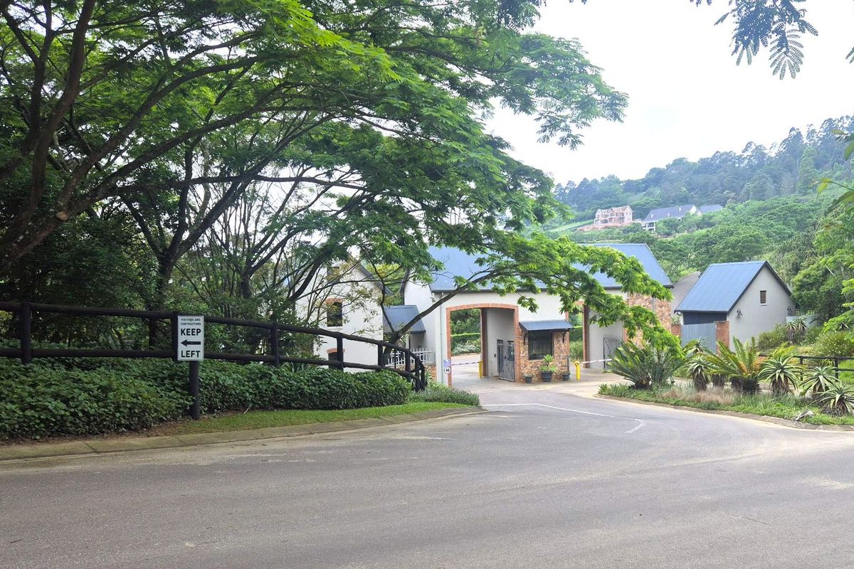 A view of the secure gated entrance to the residential estate, featuring a paved road, established trees, and a well-maintained security gatehouse.