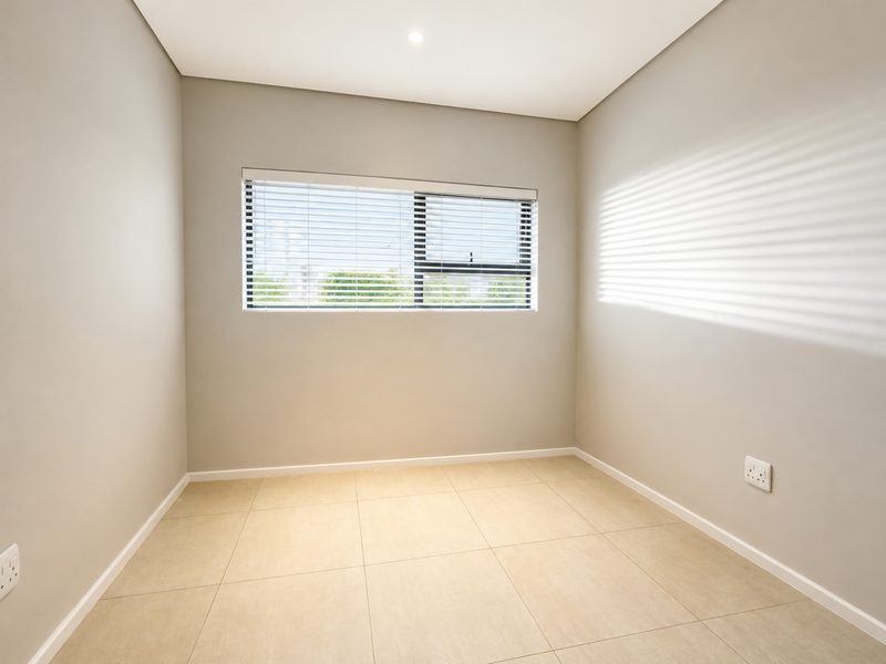 A bright, empty bedroom featuring neutral tiled flooring, clean walls, and a window fitted with venetian blinds.