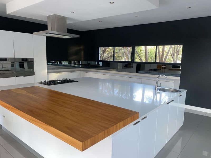 Modern kitchen featuring a large island with a timber breakfast bar, integrated gas hob, stainless steel sink, and sleek white cabinetry against a dark wall.
