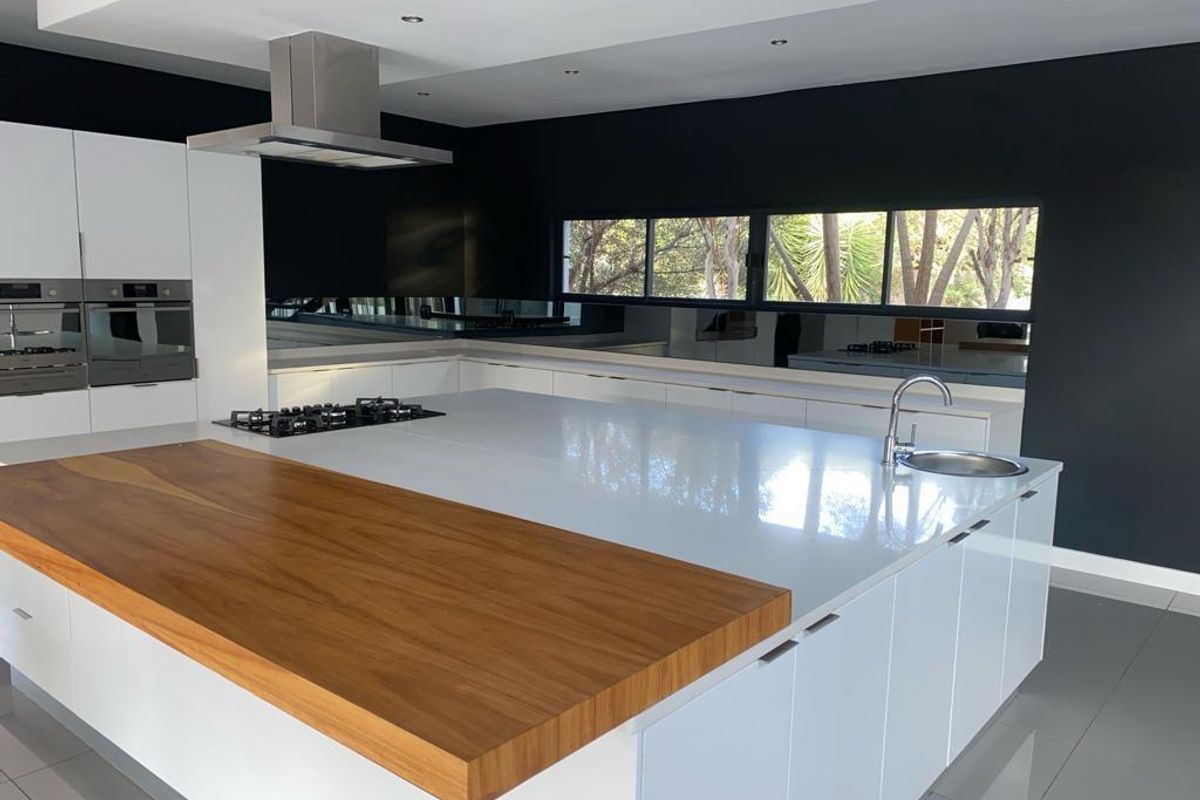 Modern kitchen featuring a large island with a timber breakfast bar, integrated gas hob, stainless steel sink, and sleek white cabinetry against a dark wall.