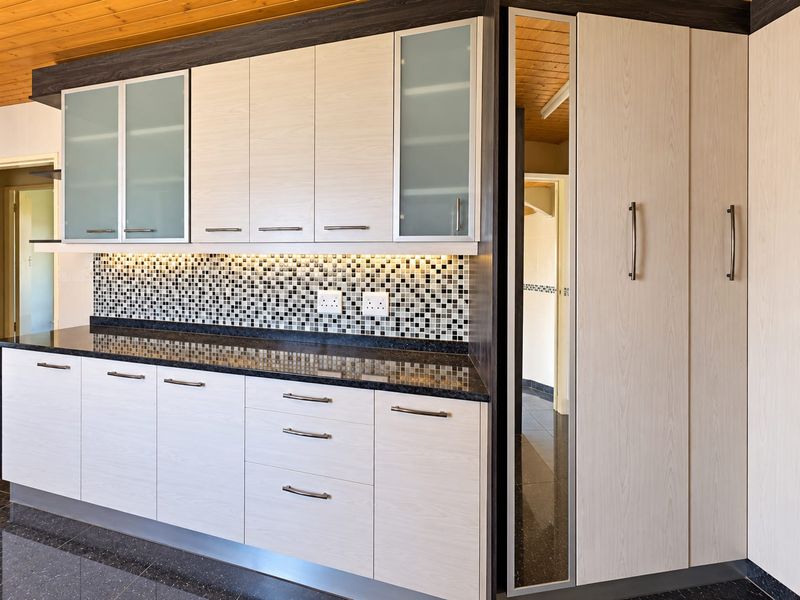 Modern kitchen featuring light wood-grain cabinetry, dark granite countertops, a mosaic tile backsplash, and integrated storage with warm under-cabinet lighting.