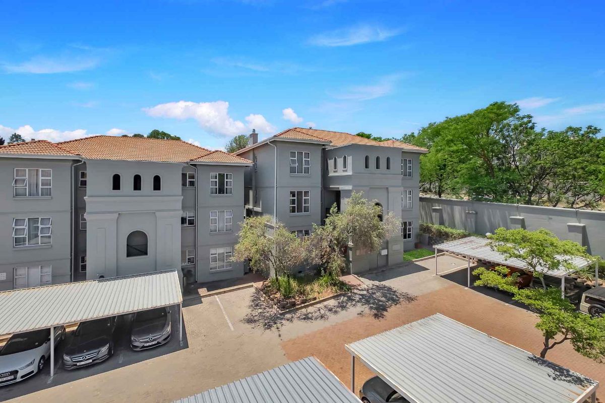 An elevated view of the apartment complex exterior, featuring grey facade, tiled roofing, and multiple covered parking bays within the paved courtyard area.