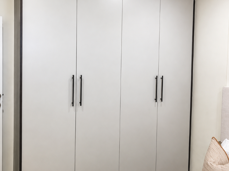 A modern bedroom featuring a large, floor-to-ceiling built-in wardrobe with sleek matte light-colored doors and dark contrasting trim.