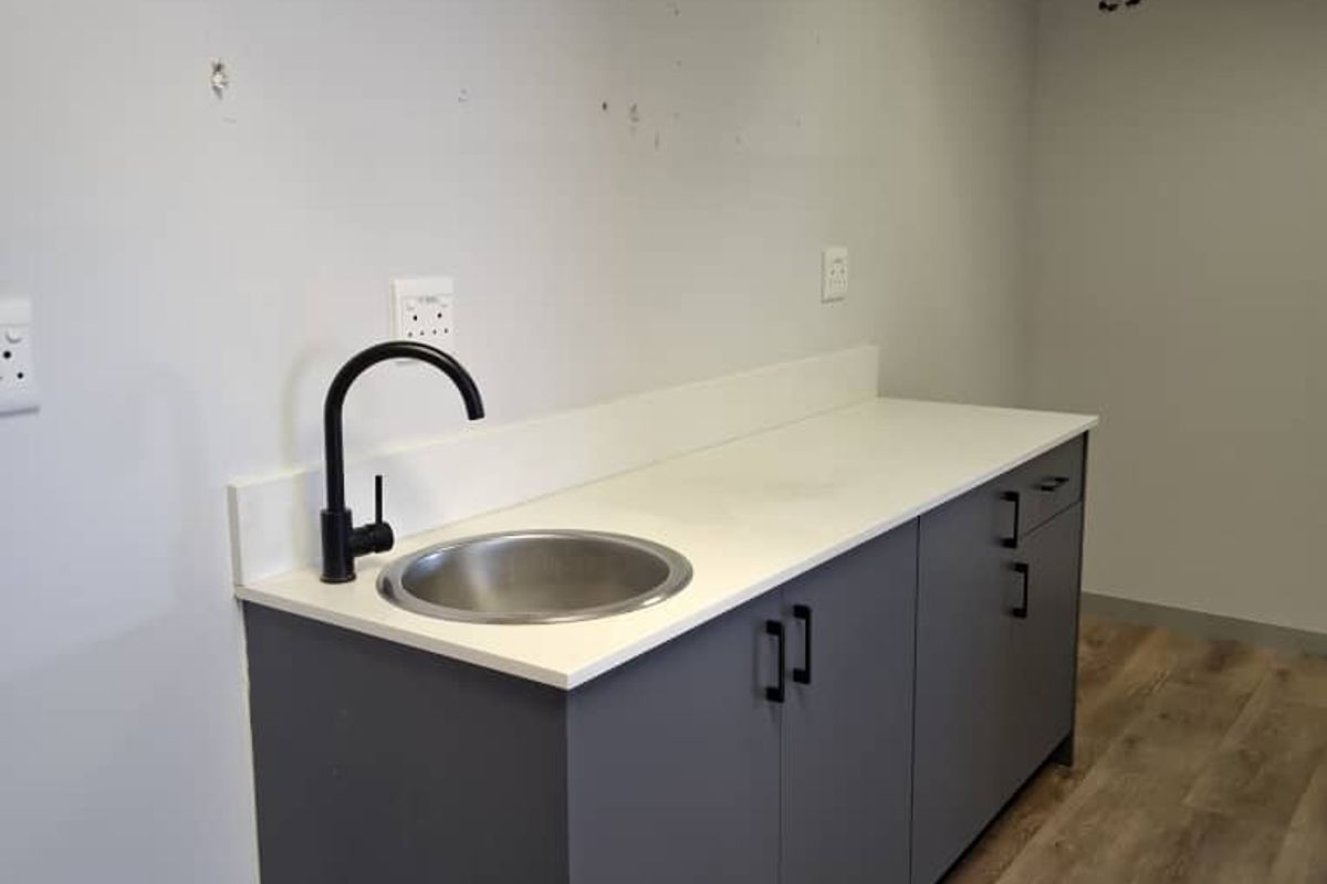 A compact office kitchenette featuring modern grey cabinetry, a stainless steel sink with a black faucet, and wood-look flooring.