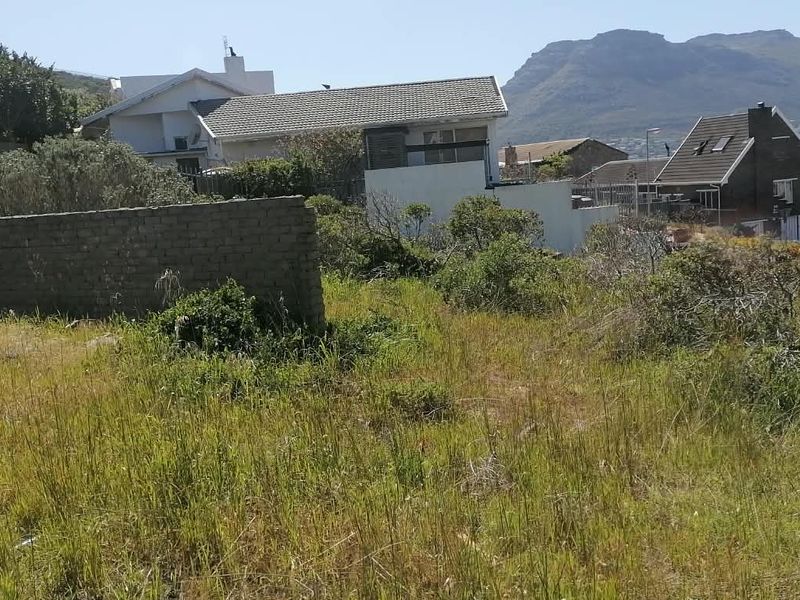 A view of a vacant residential plot featuring a partial boundary wall, surrounded by established neighborhood homes and scenic mountain views.