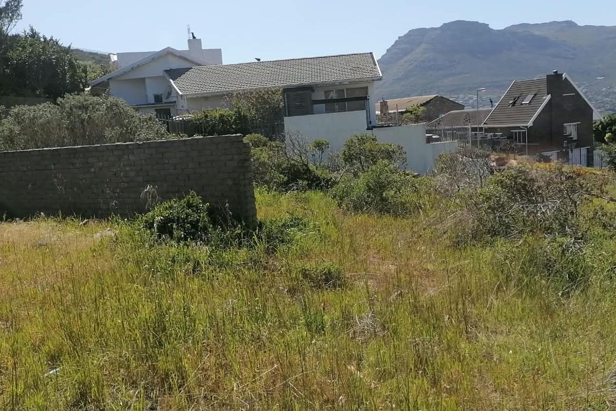 A view of a vacant residential plot featuring a partial boundary wall, surrounded by established neighborhood homes and scenic mountain views.