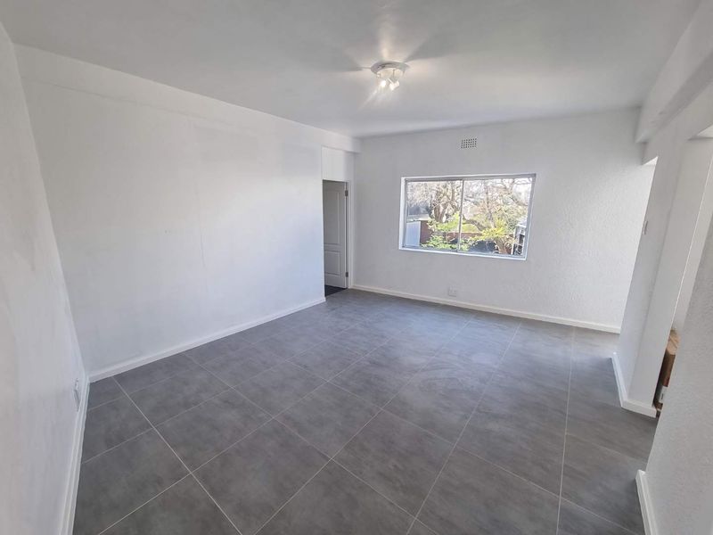 A bright, empty living room featuring neutral grey floor tiles, white walls, and a large window providing natural light.