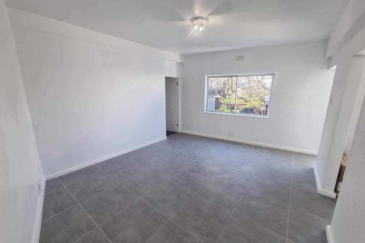 A bright, empty living room featuring neutral grey floor tiles, white walls, and a large window providing natural light.