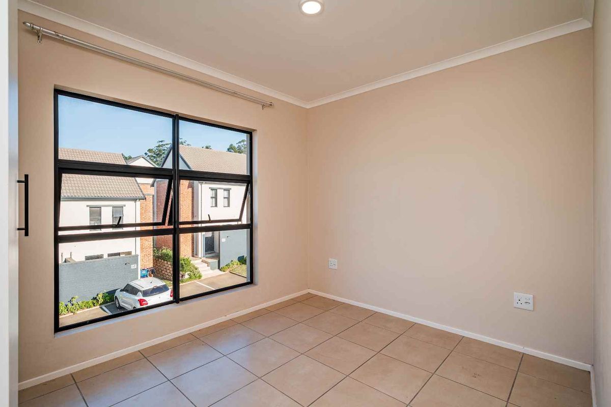 A bright, empty bedroom featuring neutral walls, tiled flooring, and a large black-framed window overlooking the complex parking area.