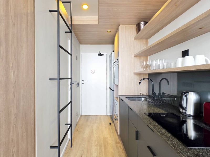 Modern galley-style kitchen featuring sleek dark cabinetry, stone countertops, integrated appliances, and warm timber ceiling accents with floating shelving.
