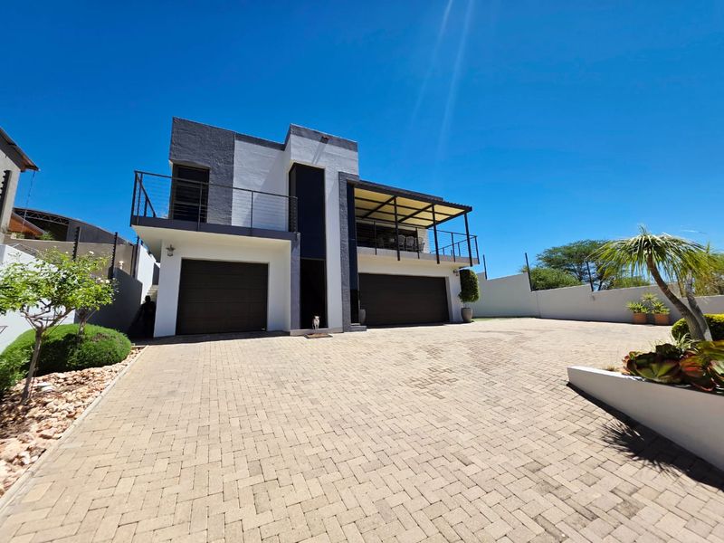 Modern double-story residence featuring a paved driveway, double garage, and an upper-level balcony with metal railings under a clear blue sky.
