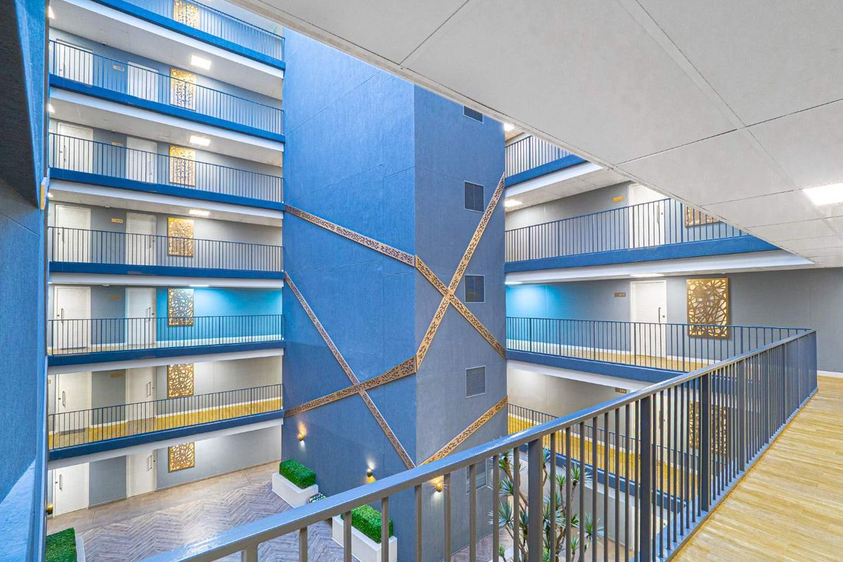 A modern apartment complex featuring a central atrium with multi-level walkways, decorative blue facade elements, and secure metal railings.