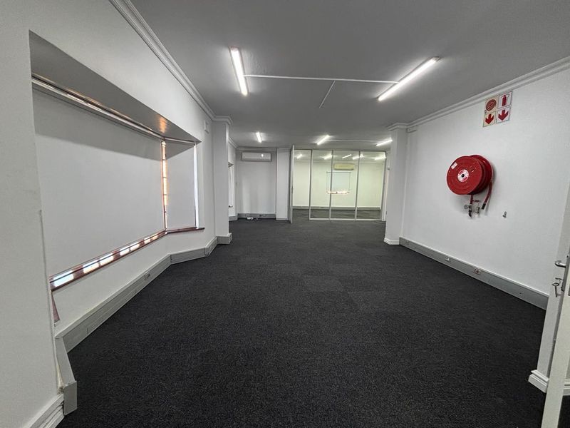 A spacious, empty office area featuring dark carpeting, white walls, a fire hose reel, and a glass-partitioned secondary workspace.