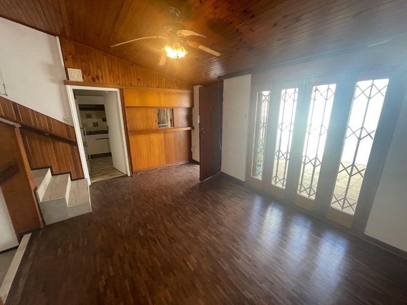 An empty living room featuring timber-paneled ceilings and walls, laminate flooring, and access to an outdoor area through security-gated doors.