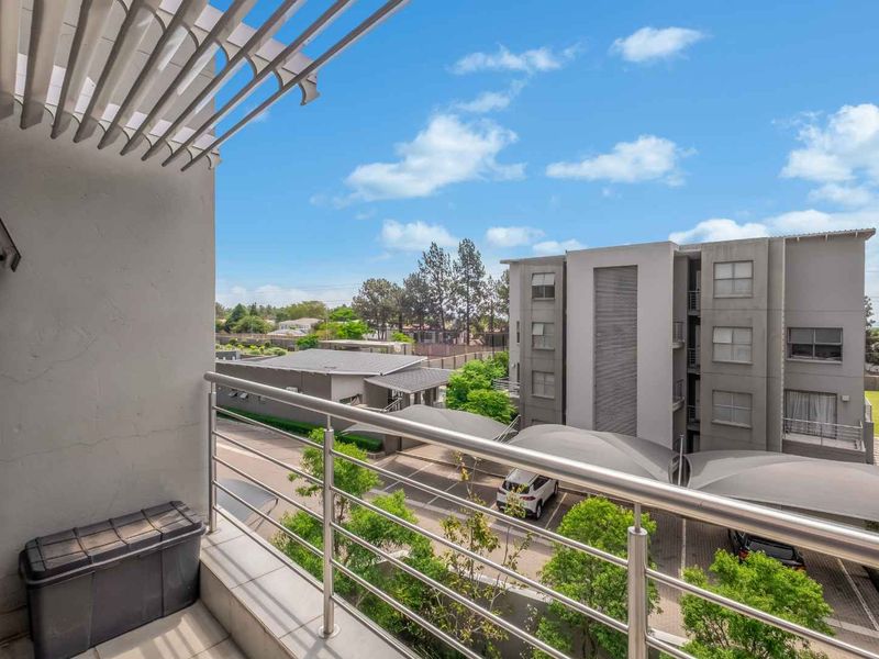 A private balcony featuring tiled flooring, a stainless steel balustrade, and an overhead pergola, offering views over the apartment complex parking area.