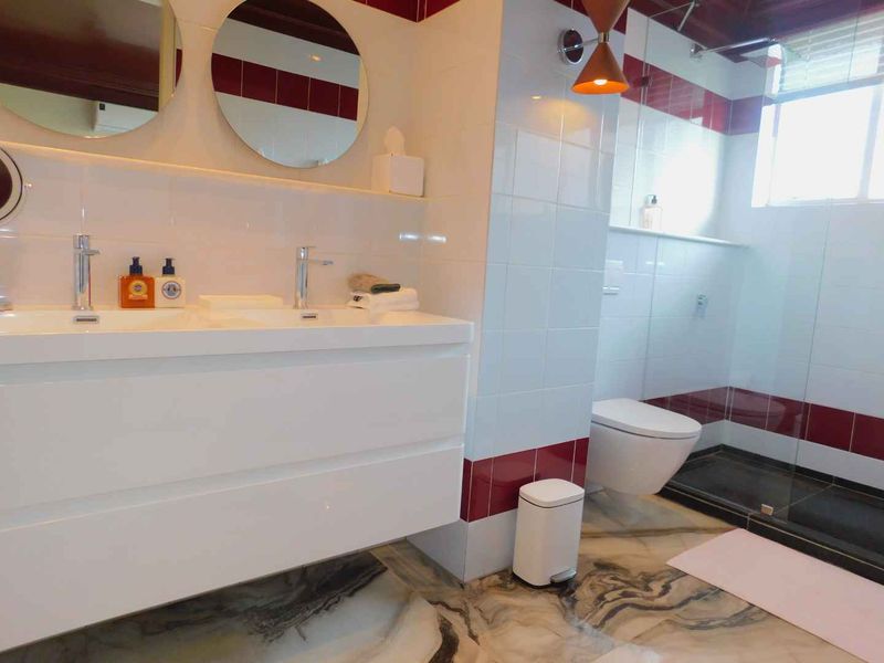 Modern bathroom featuring a double vanity, wall-mounted toilet, glass-enclosed shower, and stylish marble-effect flooring with contrasting red and white wall tiles.