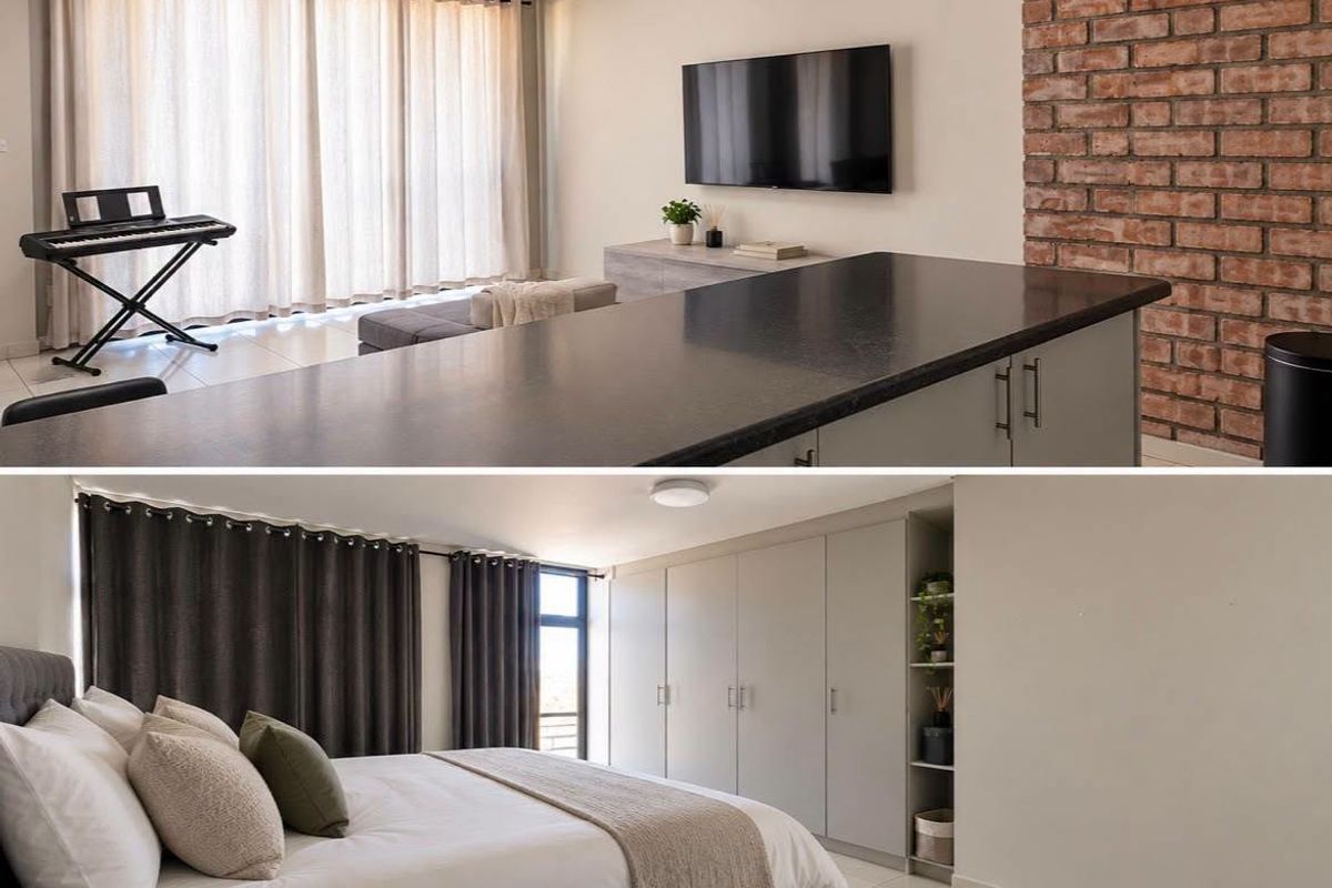 A modern open-plan living area featuring a kitchen island, exposed brick wall, and a bedroom with extensive built-in cabinetry and neutral finishes.