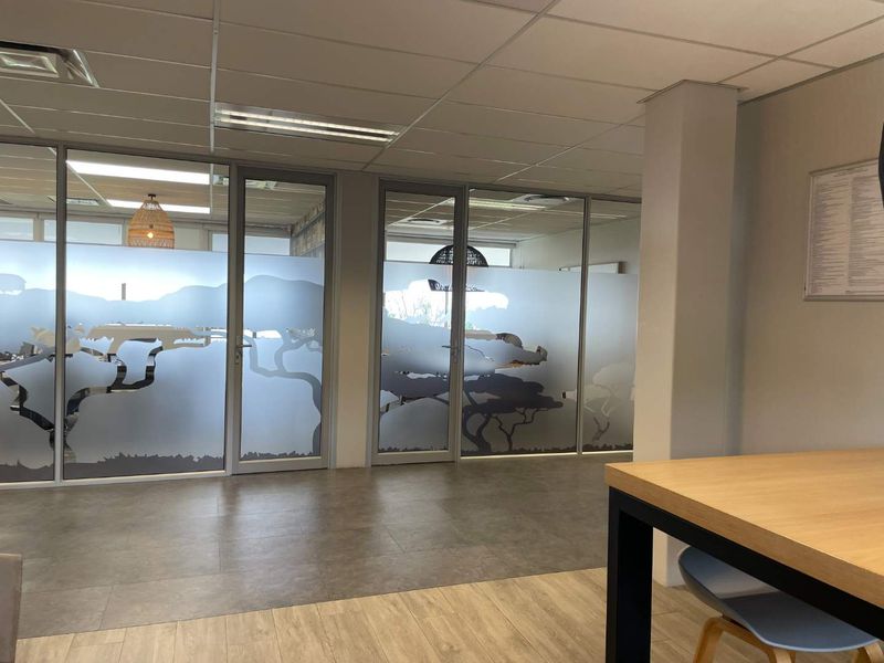 Modern office space featuring glass partition walls with decorative frosted film, suspended ceilings, and a combination of tiled and laminate flooring.