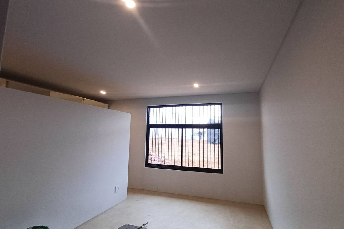 A clean, neutral-toned bedroom featuring recessed ceiling lighting and a large window fitted with security bars, offering a bright and minimalist living space.