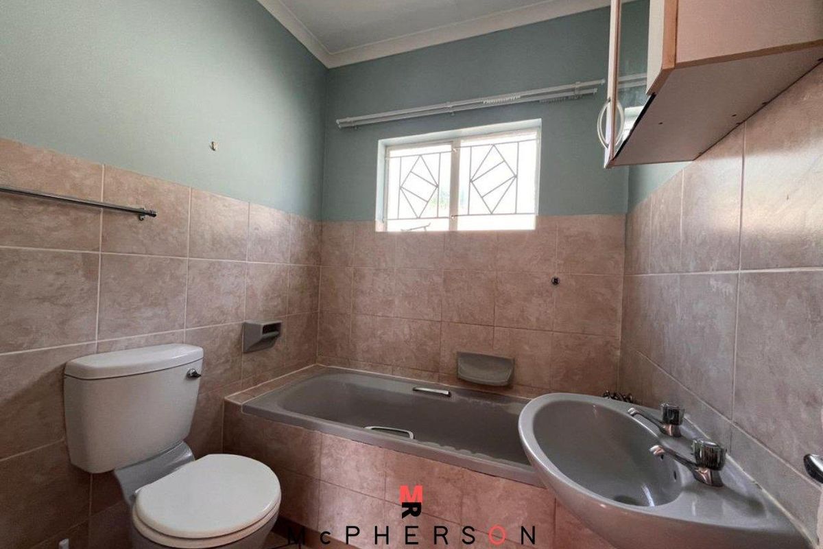 A functional bathroom featuring a built-in bathtub, pedestal basin, and toilet with neutral wall tiling and security bars on the window.