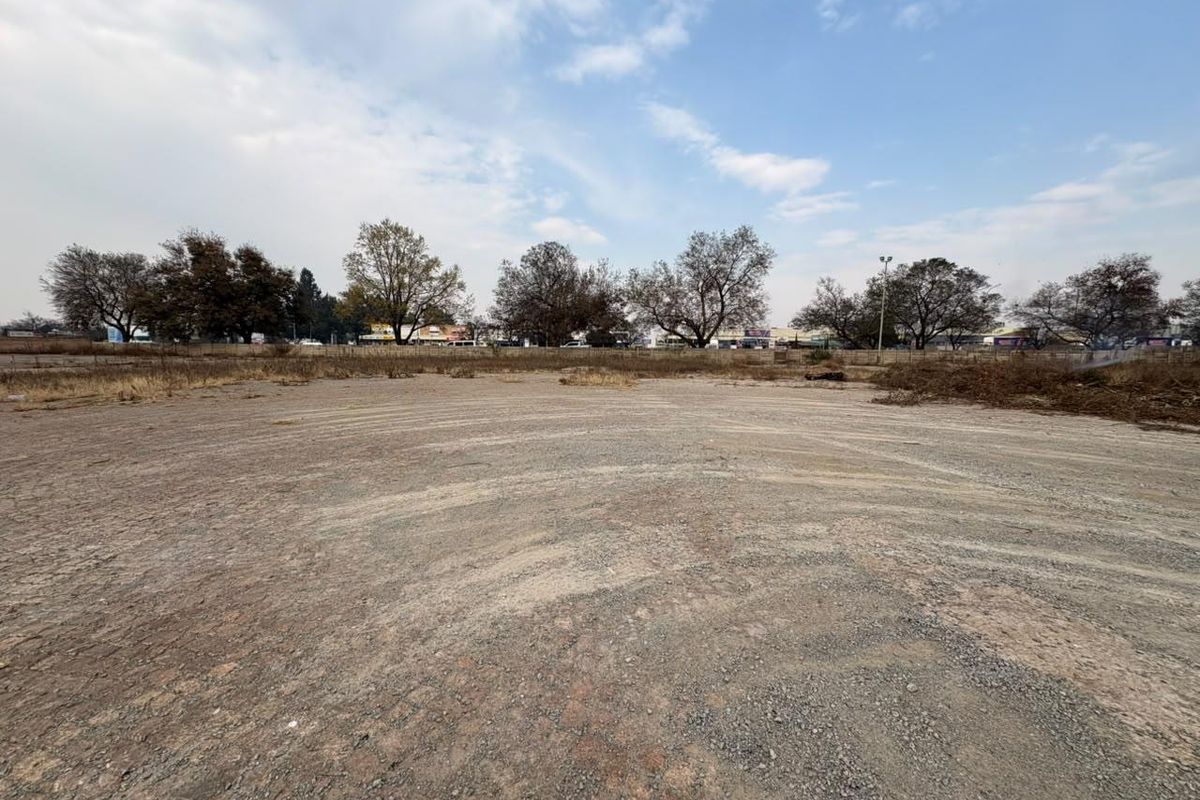 A wide-angle view of a vacant commercial land plot featuring cleared gravel terrain, perimeter fencing, and mature trees in the background.