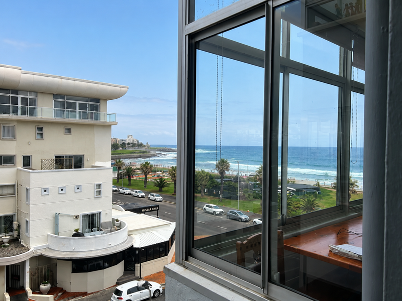 A clear view of the coastline and ocean from an elevated apartment window, overlooking the street and nearby buildings.