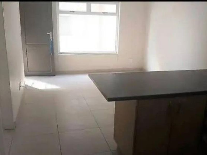 An empty open-plan living and kitchen area featuring tiled flooring, a kitchen island, and natural light from a large window.
