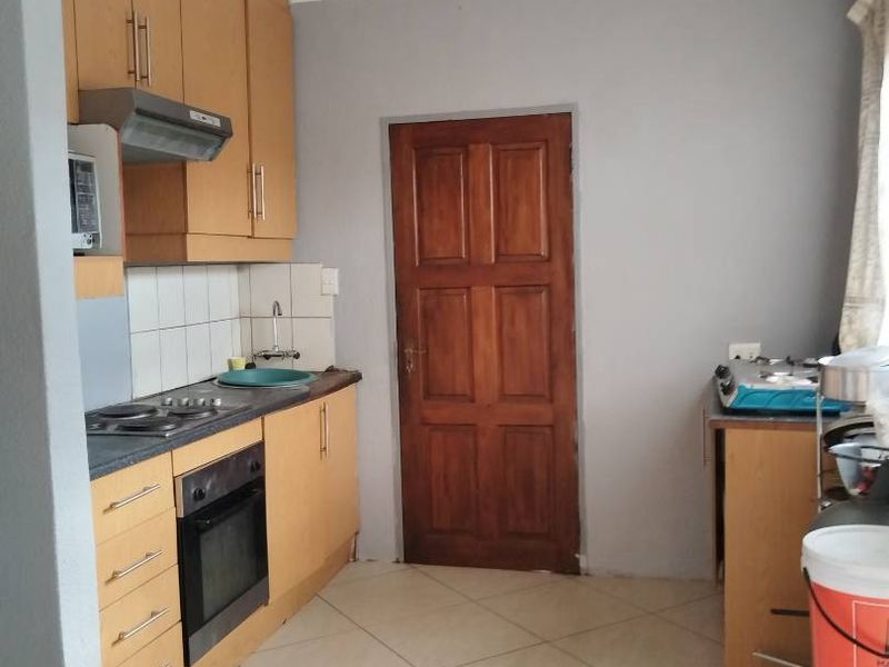A functional kitchen featuring light wood cabinetry, built-in oven and hob, tiled backsplash, and neutral floor tiles with a solid wood door.