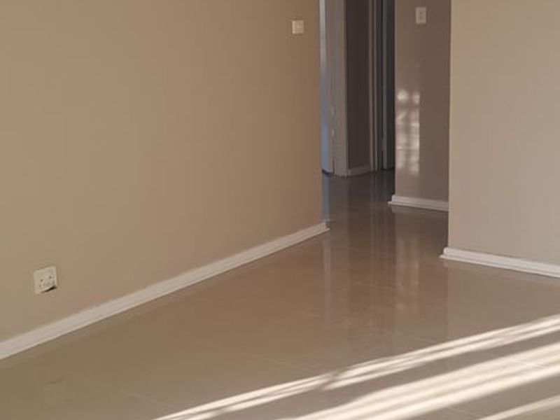A bright, empty living area featuring neutral-toned walls and polished tiled flooring, leading into a hallway with access to additional rooms.