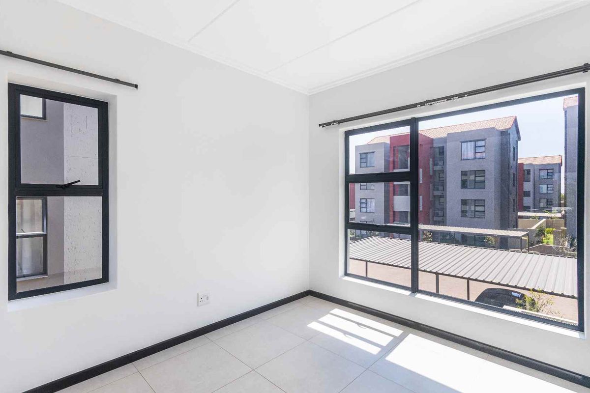 A bright, empty bedroom featuring large windows with black frames, tiled flooring, and neutral white walls.