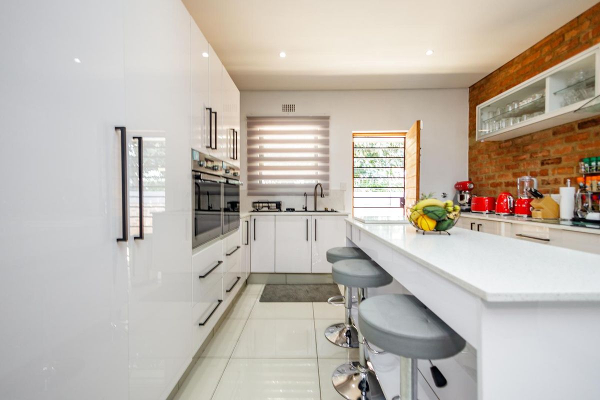 Modern kitchen featuring high-gloss white cabinetry, stone countertops, a central breakfast island, built-in double ovens, and a distinctive exposed brick feature wall.