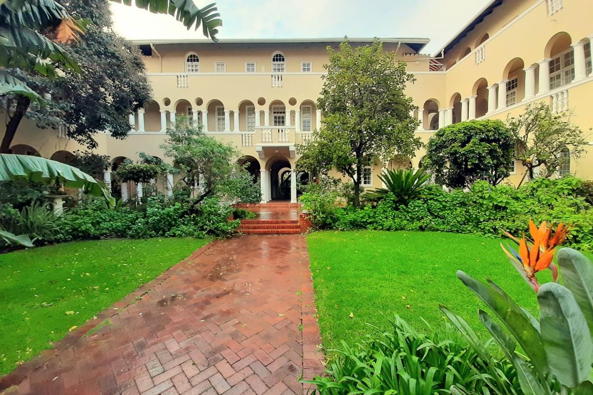 A well-maintained apartment complex courtyard featuring a brick pathway, lush garden, and classic architectural arched colonnades.