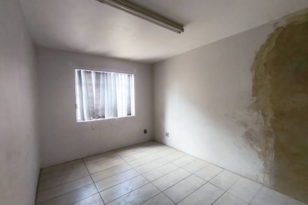 An empty bedroom featuring tiled flooring, a window with security bars, and a fluorescent light fixture, requiring wall repairs and cosmetic updates.
