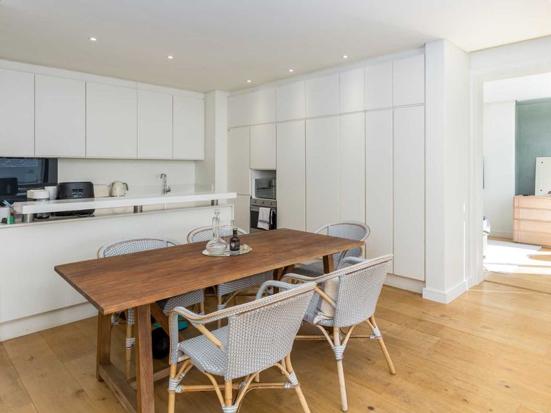 Modern open-plan kitchen and dining area featuring sleek white cabinetry, integrated appliances, stone countertops, and warm hardwood flooring.