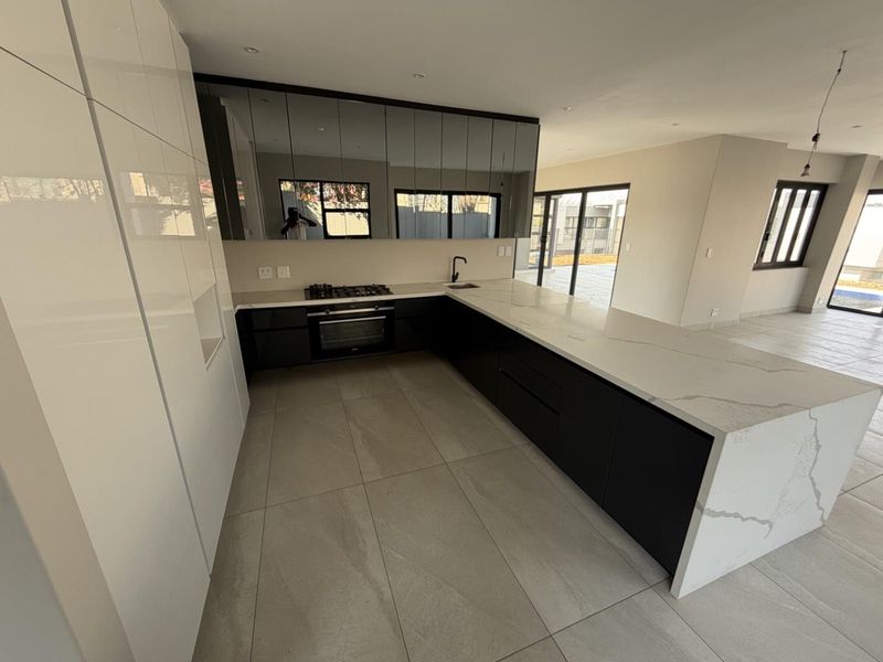 Modern open-plan kitchen featuring a large stone island, sleek dark cabinetry, integrated appliances, and a mirrored splashback, flowing into a bright living area.