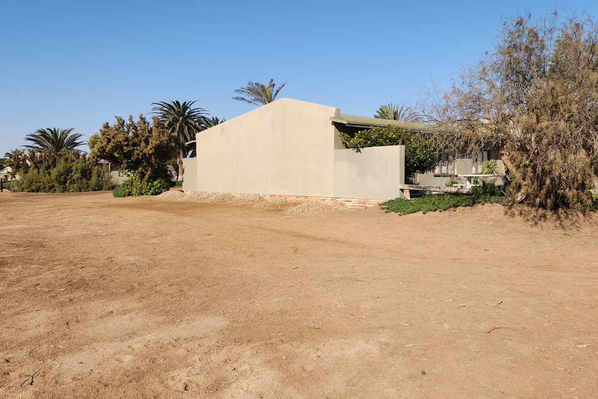 A view of the side elevation of a residential property situated on a large, undeveloped sandy plot with established trees and shrubs.