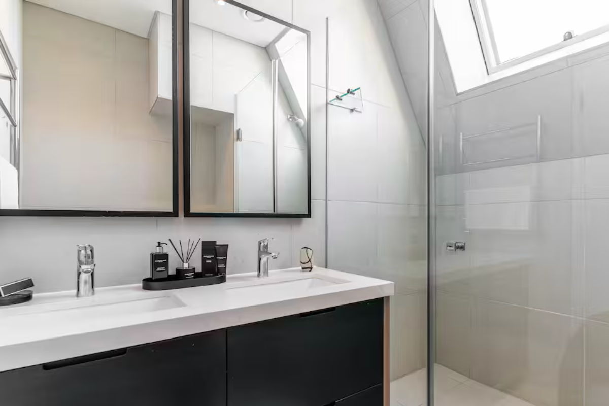 Modern bathroom featuring a sleek double vanity with integrated sinks, large mirrors, and a glass-enclosed shower under a bright skylight.