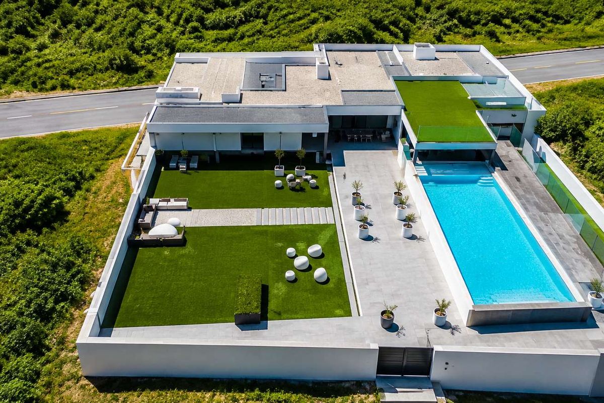 Aerial view of a modern property featuring a large swimming pool, expansive paved patio, artificial lawn, and secure boundary walls.